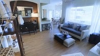 This single-floor residence showcases a thoughtfully designed open-concept layout that connects the living, dining, and kitchen areas into one cohesive, flowing space. Warm-toned hardwood flooring runs throughout, lending a sense of continuity and warmth across every zone.

The living area is anchored by a comfortable gray sofa accented with decorative blue-patterned throw pillows, complemented by a dark wood coffee table and a matching upholstered ottoman. A bold accent wall in deep brown creates a striking focal point, framing a decorative mirror and a well-appointed sideboard with storage.

The kitchen bar counter separates the living space from the dining area, with tall bar stools providing casual seating. The dining area beyond features a modern table and chairs bathed in natural light streaming through large windows dressed in sheer white curtains, creating an airy, inviting atmosphere.

Track lighting runs along the ceiling in the dining zone, adding functional illumination while reinforcing the modern aesthetic. Potted greenery and thoughtful decorative accents are placed throughout, adding life and personality to the space.

Overall, this home offers a bright, well-organized, and stylishly furnished interior that balances comfort with contemporary design, making it an ideal space for both everyday living and entertaining.