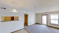 This single-floor residence offers a clean, functional layout designed for comfortable everyday living. The main living area is finished with plush wall-to-wall carpeting, which transitions to tile flooring near the entryway and kitchen pass-through zone, creating a natural delineation between spaces. Crisp white walls and trim throughout the home lend a bright, airy feel, further enhanced by natural light streaming in through a sliding glass door or large exterior window that opens to an outdoor area. A pendant light fixture adds a subtle design touch above the kitchen pass-through, which features a convenient window-style opening connecting the kitchen to the living space. Warm wood-toned bi-fold closet doors provide generous storage, adding a classic residential character to the interior. Baseboard heating units are positioned along the walls, ensuring year-round comfort. Ceiling-mounted HVAC vents contribute to efficient climate control throughout the space. The overall ambiance is neutral and move-in ready, offering a versatile canvas that adapts easily to a variety of furnishing styles and personal tastes.