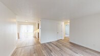 Located at 19605 SW Boones Ferry Rd in Tualatin, Oregon, this single-floor unit offers a clean, updated interior designed for comfortable everyday living. The open layout connects the living area to the rest of the unit seamlessly, with light wood-style flooring extending throughout the main living spaces, contributing to a bright and cohesive aesthetic.

The living area is generously proportioned, featuring white walls, a textured ceiling, and large vertical-blind-covered sliding glass doors that allow abundant natural light to filter in while providing access to an outdoor area. The open sightlines create a spacious feel, with clear pathways leading to additional rooms.

The kitchen is efficiently designed and features grey shaker-style cabinetry, white countertops, a stainless steel sink, and a full suite of stainless steel appliances including a refrigerator and dishwasher. A window above the counter brings in additional natural light.

A separate bedroom offers wood-style flooring, a large window, and a closet with direct door access. The bathroom includes a vanity with a sink, mirror, and brushed nickel fixtures set against clean white walls. Overall, the unit presents a fresh, well-maintained interior throughout.