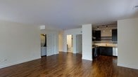 This single-floor residence offers an open, airy layout that seamlessly connects the main living area to the kitchen, creating a spacious and cohesive living environment. Rich, dark hardwood floors extend throughout, adding warmth and a sense of continuity to the space.

The kitchen is outfitted with sleek dark cabinetry that reaches toward the ceiling, providing ample storage while lending a modern, sophisticated aesthetic. A stainless steel range anchors the cooking area, complemented by tile backsplash details and track lighting overhead that ensures the workspace is well-illuminated.

The expansive main living area is defined by clean white walls and smooth ceilings, allowing for flexible furniture arrangements and an uncluttered atmosphere. Natural light filters through, enhancing the open feel of the space.

A hallway visible from the main living area leads to additional rooms, including a bathroom featuring a toilet and clean, neutral finishes. A dedicated laundry area with washer and dryer hookups adds a layer of everyday convenience. The layout is thoughtfully designed to balance open communal spaces with more private functional areas, making this a practical and well-appointed single-level home.