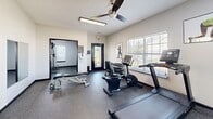 Located at 771 E Butler Rd in Mauldin, South Carolina, this fitness center offers a compact yet comprehensive workout facility spread across two exercise rooms on a single floor. The space is designed with functionality and comfort in mind, featuring durable speckled rubber flooring throughout that provides both safety and resilience underfoot.

The main exercise room is bright and welcoming, with large windows that invite ample natural light and offer outdoor views. A ceiling fan overhead ensures comfortable air circulation during workouts. Cardio equipment includes a treadmill, elliptical, and stationary bikes from TRUE Fitness, while a wall-mounted flat-screen TV adds entertainment. A wall-mounted rack holds an assortment of stability balls and resistance bands, catering to a variety of training styles.

The secondary room is dedicated to strength training, housing a multi-function cable machine, an adjustable weight bench, and a tiered dumbbell rack with a full range of free weights. Motivational fitness artwork decorates the walls, contributing to an energizing atmosphere. A glass-panel door connects the two rooms and provides direct outdoor access, further enhancing ventilation and ease of entry. A hand sanitizer station and posted fitness guidelines reflect a commitment to cleanliness and user safety throughout the facility.