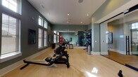 Located at 15155 Ollie Dr in Charlotte, NC, this well-appointed fitness center offers a comprehensive workout environment designed for a wide range of exercise routines. The space is finished with light hardwood-style flooring and painted in a calming sage green palette, creating an energizing yet focused atmosphere throughout.

The main workout area is generously sized and lined with large, floor-to-ceiling mirrors along one wall, visually expanding the space and supporting proper form during training. Multiple Matrix-brand cardio machines, including rowing machines and stationary bikes, are arranged in a row along the windowed wall, allowing users to exercise while benefiting from natural light streaming through a series of tall, double-pane windows. Elevated transom windows further enhance the bright and airy feel of the room.

A dedicated free weights and functional training zone features a multi-tiered rack stocked with medicine balls, kettlebells, and resistance accessories. Stability balls and a Matrix adjustable bench round out the strength training offerings. A ceiling fan overhead supports air circulation during intense workouts.

A separate, glass-walled studio room adjoins the main floor via a sleek sliding glass door, providing an ideal space for stretching, yoga, or small group training sessions. Recessed lighting throughout ensures consistent illumination regardless of the time of day, making this fitness center a fully equipped and inviting space for residents.