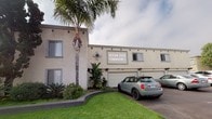 Ocean View Townhomes is a well-maintained residential complex situated in a coastal Southern California neighborhood, characterized by its classic stucco exterior finished in a neutral tone with decorative diamond accents and traditional architectural detailing. The property is single-level and presents a clean, structured façade with multiple white panel garage doors framed in gray trim, providing convenient private parking for residents. The driveway area is fully paved and accommodates multiple vehicles, offering practical and accessible parking directly adjacent to each unit. Lush, manicured green lawn areas and mature tropical landscaping — including tall palm trees and broad-leafed plants — frame the exterior, lending a warm, resort-like ambiance to the property. The surrounding neighborhood is residential in character, with tree-lined streets and open skies typical of a Southern California coastal community. A prominent building sign reading "Ocean View Townhomes" is displayed on the front facade, anchoring the property's identity. The overall exterior presents a tidy, functional, and welcoming appearance suited to the relaxed coastal lifestyle of the region.