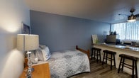 This single-floor space offers a compact yet thoughtfully arranged layout designed for comfort and functionality. The room is defined by its cool blue-grey painted walls, which lend a calm and serene ambiance throughout. Hardwood-style flooring runs the length of the room, adding warmth and a clean, modern aesthetic that contrasts nicely with the soft wall tones. The sleeping area is furnished with a full-sized bed dressed in a patterned quilt with coordinating decorative pillows, positioned against the accent wall for a clean, centered look. A wooden bedside table with a classic lamp provides practical bedside lighting. Along the opposite wall, a narrow wooden counter serves as a workspace or vanity area, accompanied by two black bar-height stools — a versatile touch that maximizes the use of space. A small framed piece of art and a few decorative accents on the counter add a personal, homey feel. The ceiling is clean and unadorned, contributing to the room's understated simplicity. Overall, the space presents a cozy, well-organized environment with a cohesive color palette and practical furnishings suited for everyday living.