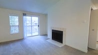This single-floor space centers around a bright, open living area designed for comfort and functionality. The room is finished with wall-to-wall carpeting in a neutral tone, providing a soft, cohesive foundation throughout. A standout feature is the wood-burning fireplace, framed by a clean white tile surround and matching tiled hearth, which serves as a natural focal point along one wall. The fireplace adds both visual interest and practical warmth to the living space.

Natural light is a notable characteristic of this space, with a large sliding glass door along one wall drawing in abundant daylight and offering views of a neighboring exterior streetscape. The sliding door also provides direct access to a private balcony enclosed by a white railing, extending the living area outdoors. The walls are finished in a soft, neutral white, complementing the light tones of the tile and carpet. The ceiling features a subtle textured finish, adding modest architectural detail. Overall, the space presents a clean, functional layout with a warm ambiance, well-suited for everyday living.
