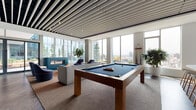 Located at 230 E Burnside Street in Portland's vibrant inner eastside, this contemporary amenity lounge offers a thoughtfully designed social space with an open, airy layout bathed in natural light. Floor-to-ceiling windows line the perimeter, framing panoramic views of the Portland skyline and surrounding hills. A striking linear slatted ceiling adds architectural depth and visual rhythm throughout the space.

The main lounge area features rich dark hardwood flooring and a curated mix of modern furnishings, including swivel lounge chairs with bold accent trim, modular sofas, and sculptural side tables arranged across layered area rugs. A full-size pool table anchors the central gathering zone, complemented by a sleek white wet bar with bar seating and a large mounted display screen — ideal for entertainment and social events.

A dedicated media nook, accented by a calming blue feature wall, provides a more intimate setting with rounded upholstered chairs arranged around a large wall-mounted screen. Adjacent to the main lounge, a compact meeting or dining area offers a streamlined white table with Scandinavian-style stools positioned beside bright corner windows. Direct access to an outdoor terrace further extends the social space, enhancing the connection between interior comfort and the dynamic Portland cityscape beyond.