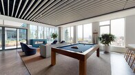 Located at 230 E Burnside Street in Portland's vibrant inner eastside, this contemporary amenity lounge offers a thoughtfully designed social space with an open, airy layout bathed in natural light. Floor-to-ceiling windows line the perimeter, framing panoramic views of the Portland skyline and surrounding hills. A striking linear slatted ceiling adds architectural depth and visual rhythm throughout the space.

The main lounge area features rich dark hardwood flooring and a curated mix of modern furnishings, including swivel lounge chairs with bold accent trim, modular sofas, and sculptural side tables arranged across layered area rugs. A full-size pool table anchors the central gathering zone, complemented by a sleek white wet bar with bar seating and a large mounted display screen — ideal for entertainment and social events.

A dedicated media nook, accented by a calming blue feature wall, provides a more intimate setting with rounded upholstered chairs arranged around a large wall-mounted screen. Adjacent to the main lounge, a compact meeting or dining area offers a streamlined white table with Scandinavian-style stools positioned beside bright corner windows. Direct access to an outdoor terrace further extends the social space, enhancing the connection between interior comfort and the dynamic Portland cityscape beyond.