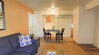 This single-floor residential unit offers a practical and comfortable layout designed to maximize everyday living. The open-concept main area combines the living room and dining space in a seamless, flowing arrangement. The living area features warm, neutral-toned carpeting and is accented by a bold orange feature wall that adds a pop of color and warmth to the space. A plush blue sofa anchors the living area, complemented by a coffee table for a welcoming, relaxed atmosphere. Adjacent to the living room, the dining area accommodates a bistro-style high-top table with matching chairs, making it ideal for casual meals and entertaining. The kitchen is partially open to the main living space, offering visibility and connectivity. It features warm honey-toned wood cabinetry, a range, and functional countertop space. White interior doors and neutral cream-colored walls throughout the unit contribute to a bright and clean aesthetic. Ceiling-mounted lighting provides consistent illumination across the main living areas. The layout is thoughtfully designed for efficient use of space, making it a comfortable and inviting place to call home.