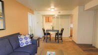 This single-floor residential unit offers a practical and comfortable layout designed to maximize everyday living. The open-concept main area combines the living room and dining space in a seamless, flowing arrangement. The living area features warm, neutral-toned carpeting and is accented by a bold orange feature wall that adds a pop of color and warmth to the space. A plush blue sofa anchors the living area, complemented by a coffee table for a welcoming, relaxed atmosphere. Adjacent to the living room, the dining area accommodates a bistro-style high-top table with matching chairs, making it ideal for casual meals and entertaining. The kitchen is partially open to the main living space, offering visibility and connectivity. It features warm honey-toned wood cabinetry, a range, and functional countertop space. White interior doors and neutral cream-colored walls throughout the unit contribute to a bright and clean aesthetic. Ceiling-mounted lighting provides consistent illumination across the main living areas. The layout is thoughtfully designed for efficient use of space, making it a comfortable and inviting place to call home.