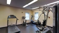 This single-floor fitness room offers a practical and well-organized workout space designed to accommodate a range of exercise needs. The room features a rubber-floored exercise area that provides both safety and durability underfoot, ideal for high-impact activities. Equipment includes a treadmill, an upright stationary bike, an elliptical trainer, and a multi-function cable resistance machine, offering users options for both cardio and strength training. Two windows along the back wall allow natural light to enter the space, contributing to a more open and comfortable atmosphere. Overhead fluorescent lighting ensures the room remains well-lit throughout the day. The walls are finished in a neutral beige tone, giving the space a clean and uncluttered appearance. The layout is efficient, with equipment arranged to allow reasonable movement between machines. The dark rubber flooring contrasts with the light walls, adding a functional aesthetic suited to a fitness environment. Overall, the space is straightforward and purpose-driven, providing the essentials for a productive workout routine.