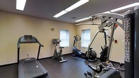 This single-floor fitness room offers a practical and well-organized workout space designed to accommodate a range of exercise needs. The room features a rubber-floored exercise area that provides both safety and durability underfoot, ideal for high-impact activities. Equipment includes a treadmill, an upright stationary bike, an elliptical trainer, and a multi-function cable resistance machine, offering users options for both cardio and strength training. Two windows along the back wall allow natural light to enter the space, contributing to a more open and comfortable atmosphere. Overhead fluorescent lighting ensures the room remains well-lit throughout the day. The walls are finished in a neutral beige tone, giving the space a clean and uncluttered appearance. The layout is efficient, with equipment arranged to allow reasonable movement between machines. The dark rubber flooring contrasts with the light walls, adding a functional aesthetic suited to a fitness environment. Overall, the space is straightforward and purpose-driven, providing the essentials for a productive workout routine.