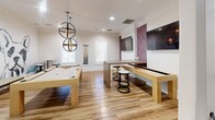 Located in Fullerton, California, this thoughtfully designed single-floor amenity space offers a diverse range of functional areas that cater to socializing, dining, working, and recreation. The layout connects multiple hallways, two living areas, a dining room, and a private office, creating a seamless flow throughout.

The welcoming lounge area features elegant marble-style tile flooring, a cream sofa accented with soft pink pillows, and delicate textured wallpaper, setting a refined yet comfortable tone. Nearby, a compact lounge nook provides a more intimate setting with a round table, upholstered stools, curated wall art, and a mounted flat-screen TV.

The bright dining area is anchored by warm hardwood-style flooring, a central dining table with vibrant chartreuse chairs, a bar-height counter with patterned seating, and a trio of modern pendant lights. Natural light pours in through large windows, enhancing the clean, contemporary aesthetic. A bold "eat" wall sign adds a playful touch.

The game room is a standout, featuring a full-size pool table and a shuffleboard table beneath striking orb-style chandeliers, with a mounted TV against a herringbone-patterned accent wall. The private office station offers a dedicated computer workstation with a sleek pendant light and ample natural light from surrounding windows. Throughout, crown molding and recessed lighting add a polished finish.