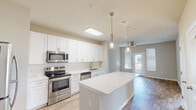 This single-floor residence is designed with a clean, modern aesthetic that emphasizes an open and functional layout. The kitchen serves as the centerpiece of the living space, featuring an expansive white center island topped with a smooth white countertop, illuminated by elegant pendant lights that hang at varying heights. Crisp white shaker-style cabinetry lines the walls, paired with white subway tile backsplash and complemented by a full suite of stainless steel appliances, including a range, over-the-range microwave, dishwasher, and refrigerator. A undermount sink with a brushed nickel faucet adds a refined touch.

The kitchen flows seamlessly into the living area, where light wood-style flooring extends throughout, creating a warm and cohesive feel. A ceiling fan provides comfort in the main living space, which receives natural light through windows fitted with horizontal blinds and a glass-paneled exterior door. Additional rooms feature the same wood-style flooring and ceiling fans.

The bathroom presents white cabinetry, a white quartz-style countertop, an undermount sink with brushed nickel hardware, and a large wall mirror. The overall palette of warm neutral tones and white finishes gives the space a bright, welcoming ambiance.