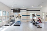 Located at 4659 Dawson Blvd in Atlanta, GA, this fitness center offers a well-equipped, single-floor space designed to support a comprehensive workout experience. The interior gym features a clean, modern aesthetic with bright white walls accented by bold horizontal navy and light blue stripes, creating a vibrant yet focused atmosphere. Recessed lighting and multiple windows allow natural light to fill the space, offering views of the surrounding outdoor area and pool. The patterned geometric flooring adds a contemporary touch throughout.

The cardio section is outfitted with treadmills, an elliptical trainer, and a stationary spin bike, complemented by a wall-mounted flat-screen TV for entertainment during workouts. The strength training area includes a weight bench, multi-use cable machine, medicine ball rack, and free weights, providing a well-rounded selection of equipment. A large closet offers additional storage, and support columns subtly define different workout zones without disrupting the open layout.

Extending the fitness experience outdoors, an adjacent turf exercise area features pull-up bars and functional training equipment, enclosed by iron fencing and surrounded by mature trees that provide natural shade. The combination of thoughtfully designed indoor facilities and an accessible outdoor training area makes this a comprehensive fitness destination in the Doraville area of Metro Atlanta.