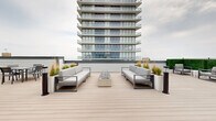 Located on the 4th floor of a modern high-rise in Asbury Park, NJ, Sunset Terrace is a thoughtfully designed rooftop outdoor space that offers a variety of amenities for relaxation and social gathering. The terrace is finished with light-toned composite decking that runs throughout, providing a clean, cohesive aesthetic across the entire level.

At the heart of the space is a central lounge area featuring two large, contemporary sofas with plush grey cushions, flanking a sleek elongated fire table filled with decorative river stones — a striking focal point ideal for gathering. Adjacent to this primary seating area, a dedicated dining zone accommodates multiple table-and-chair sets, perfect for al fresco meals while taking in open sky views of the Asbury Park skyline.

A private cabana with a retractable canopy and blue cushioned daybed offers a shaded retreat, separated from the main areas by tall white planters filled with ornamental grasses that add a natural, textural element to the design. Additional lounge seating with modular furniture and a side table is tucked nearby, creating an intimate secondary gathering zone.

Lush green hedge panels line portions of the perimeter, contrasting with the clean grey and white palette of the surrounding architecture. The rooftop is easily accessed via glass double doors leading to the building's interior, ensuring seamless indoor-outdoor connectivity throughout this well-appointed outdoor retreat.