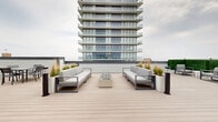 Located on the 4th floor of a modern high-rise in Asbury Park, NJ, Sunset Terrace is a thoughtfully designed rooftop outdoor space that offers a variety of amenities for relaxation and social gathering. The terrace is finished with light-toned composite decking that runs throughout, providing a clean, cohesive aesthetic across the entire level.

At the heart of the space is a central lounge area featuring two large, contemporary sofas with plush grey cushions, flanking a sleek elongated fire table filled with decorative river stones — a striking focal point ideal for gathering. Adjacent to this primary seating area, a dedicated dining zone accommodates multiple table-and-chair sets, perfect for al fresco meals while taking in open sky views of the Asbury Park skyline.

A private cabana with a retractable canopy and blue cushioned daybed offers a shaded retreat, separated from the main areas by tall white planters filled with ornamental grasses that add a natural, textural element to the design. Additional lounge seating with modular furniture and a side table is tucked nearby, creating an intimate secondary gathering zone.

Lush green hedge panels line portions of the perimeter, contrasting with the clean grey and white palette of the surrounding architecture. The rooftop is easily accessed via glass double doors leading to the building's interior, ensuring seamless indoor-outdoor connectivity throughout this well-appointed outdoor retreat.