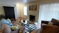 This single-floor residence offers a comfortable and well-appointed living space designed with both function and style in mind. The living room serves as the heart of the home, centered around a classic white-painted brick fireplace that adds warmth and character to the room. Hardwood-style flooring extends throughout, providing a clean and cohesive aesthetic that ties the space together.

Natural light flows generously into the living area through large windows dressed with vertical blinds, creating a bright and airy atmosphere. A sliding glass door further enhances the sense of openness while offering access to the exterior. The textured ceiling adds a traditional architectural detail that complements the overall design.

The entryway is clearly defined, featuring a solid front door that provides a welcoming transition into the home. Decorative artwork displayed above the fireplace adds a touch of personality and color to the neutral wall palette. The living room comfortably accommodates a full seating arrangement around a glass-top coffee table, making it ideal for both relaxing and entertaining. Overall, this home presents a functional, inviting layout with classic design elements that create a warm and livable environment throughout.