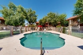 Located at 1701 N Palo Verde Dr in Goodyear, Arizona, this outdoor recreational area centers around a generously sized freeform swimming pool with clear turquoise water. The pool features gradually varying depths, entry steps, and handrail access, making it accessible and inviting under the Arizona sun. Surrounding the pool is a wide concrete sundeck outfitted with white lounge chairs and red patio umbrellas, offering both sun and shade options for relaxation. The entire pool area is enclosed by a metal security fence with a gated entry, accented by brick pillar details that complement the warm, southwestern architectural style of the adjacent buildings. A covered pavilion-style structure near the gate provides additional shade and houses posted pool rules and safety equipment, including a life ring. Lush, mature trees line the perimeter, casting dappled shade and creating a pleasant, park-like atmosphere that offers a cool contrast to the desert surroundings. The buildings visible beyond the fence feature tile rooflines, warm earth-tone stucco exteriors, and balconies with railings, consistent with classic Arizona residential architecture. The overall setting is bright, open, and well-maintained, providing a comfortable and functional outdoor amenity space suited to the warm Goodyear climate.