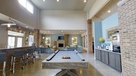 Located at 100 Comet Creek Lane in Summerville, South Carolina, this single-floor community amenity space is designed to impress with its grand, open-concept layout. Soaring ceilings amplify the sense of spaciousness, while exposed brick columns add warmth and architectural character throughout the interior.

At the heart of the space sits a full-size pool table, serving as a natural gathering point for residents. Beyond it, a generous lounge area is anchored by a clean-lined fireplace framed by brick columns, with comfortable seating arrangements ideal for conversation or relaxation. A wall-mounted television completes the entertainment setup in this central living zone.

Recessed lighting and pendant fixtures are thoughtfully distributed across the ceiling, providing layered illumination that enhances the open atmosphere. Polished concrete flooring runs throughout, lending an industrial-modern aesthetic that pairs well with the soft blue-gray wall tones.

A bar-height counter with seating offers an additional casual area for socializing, while a side counter with cabinetry — accented with decorative greenery — adds a refined, functional touch. Large windows allow natural light to filter into the space, contributing to the bright and welcoming ambiance.

Overall, this amenity space delivers a polished, well-appointed environment suited for both active recreation and leisurely gatherings.