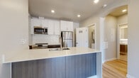 This well-appointed single-floor unit showcases a clean, contemporary design with thoughtful finishes throughout. The open-concept kitchen serves as a central highlight, featuring a large island with a light quartz countertop and textured wood-tone cabinetry. White upper cabinets with a subway tile backsplash complement a full suite of stainless steel appliances, including a refrigerator, range, and over-the-range microwave. Recessed lighting illuminates the kitchen and adjoining areas, enhancing the bright, airy atmosphere. Light hardwood-style flooring runs seamlessly throughout the unit, contributing to a cohesive and modern aesthetic. The bathroom is elegantly finished with large-format travertine-style tile flooring, a wall-mounted floating vanity with integrated sink, and a backlit frameless mirror that adds a refined touch. The living area benefits from a sliding glass door opening to a private balcony, inviting natural light into the space. Additional rooms are clean and versatile, with neutral walls, large windows fitted with horizontal blinds, and wall-mounted climate control units. An in-unit laundry area is also accessible, adding everyday convenience to this thoughtfully designed residence.
