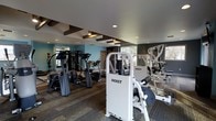 This fitness center is a thoughtfully designed workout facility offering a full range of exercise equipment for both cardio and strength training. The space is outfitted with professional-grade Hoist brand machines, including multi-functional strength stations and cable systems, providing users with a versatile and effective training environment. A lineup of cardio equipment, including elliptical trainers and treadmills, occupies a dedicated area of the floor, allowing for efficient use of the available space. The flooring features durable, textured carpet tiles suited for a high-traffic fitness environment. Large mirrored walls on one side of the room enhance the sense of openness while allowing users to monitor their form during workouts. Natural light filters in through windows, complementing the recessed ceiling lighting that illuminates the space evenly. Decorative dark wood beam accents along the ceiling add a polished, modern aesthetic against the neutral and teal-accented walls. The overall layout is open and well-organized, ensuring ease of movement between equipment stations. This fitness center is designed to deliver a functional and comfortable environment for a well-rounded workout routine.