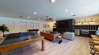 This single-floor space is thoughtfully designed as a versatile entertainment and recreation hub. The open layout is anchored by light hardwood flooring throughout, creating a cohesive flow between distinct activity zones. Recessed lighting and a brass ceiling fan illuminate the main lounge area, which features a tufted leather sofa, upholstered chairs, a wooden coffee table, and a large wall-mounted television set against a dark shiplap accent wall with a fireplace. Decorative skateboards and curated artwork add a playful, urban character to the space.

The game area is generously sized to accommodate a ping-pong table and a foosball table side by side, with ample room for play. Multiple windows along the perimeter bring in natural light, and an interior glass window panel offers a view into an adjacent fitness area. A bold pop-art mural adds a striking visual anchor near the arcade game corner.

The kitchenette is a standout feature, finished with deep navy shaker cabinets, white countertops, a stainless steel sink and appliances, and a navy star-patterned tile backsplash. A floating wood shelf above, illuminated by brass sconce lighting, completes the polished yet casual aesthetic. A counter-height bar area with seating rounds out the social amenities of this well-appointed space.