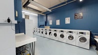 This communal laundry facility is designed with practicality and efficiency in mind. The room is defined by its bold blue accent walls, which contrast cleanly against the light-colored flooring and white equipment, creating a bright and organized atmosphere. An entire wall is lined with a row of modern front-loading washing machines and dryers, offering ample capacity for simultaneous use. The machines feature digital control panels, making them straightforward to operate. Instructional signage is posted on the walls, providing helpful guidance on laundry and drying techniques. A white folding table with hairpin legs offers a dedicated surface for sorting and folding laundry, while a wire laundry cart nearby adds further convenience. Additional storage, including a white cabinet, is available along one wall. Overhead, exposed ductwork and fluorescent lighting run across the ceiling, contributing to the utilitarian character of the space. Ventilation is supported by louvered wall vents. The overall layout is clean, spacious, and thoughtfully arranged to accommodate multiple users comfortably, making this laundry room a highly functional shared amenity.