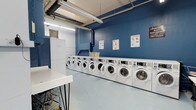 This communal laundry facility is designed with practicality and efficiency in mind. The room is defined by its bold blue accent walls, which contrast cleanly against the light-colored flooring and white equipment, creating a bright and organized atmosphere. An entire wall is lined with a row of modern front-loading washing machines and dryers, offering ample capacity for simultaneous use. The machines feature digital control panels, making them straightforward to operate. Instructional signage is posted on the walls, providing helpful guidance on laundry and drying techniques. A white folding table with hairpin legs offers a dedicated surface for sorting and folding laundry, while a wire laundry cart nearby adds further convenience. Additional storage, including a white cabinet, is available along one wall. Overhead, exposed ductwork and fluorescent lighting run across the ceiling, contributing to the utilitarian character of the space. Ventilation is supported by louvered wall vents. The overall layout is clean, spacious, and thoughtfully arranged to accommodate multiple users comfortably, making this laundry room a highly functional shared amenity.