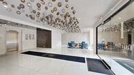 Located at 4247 Locust Street in Philadelphia's University City neighborhood, this elegantly appointed lobby serves as the welcoming centerpiece of The Fairfax. The space immediately commands attention with its most distinctive feature: a dramatic ceiling installation composed of dozens of metallic orbs in gold, silver, and bronze tones, suspended at varying heights to create a captivating, cloud-like sculptural display overhead.

The floor is finished in large-format, high-gloss white tiles that reflect light throughout the open, generously proportioned interior, enhancing the sense of brightness and spaciousness. A branded entry runner leads visitors from the glass-enclosed automatic entrance doors toward the heart of the lobby. Two separate glass entry vestibules provide access from the street, framing views of the surrounding neighborhood.

The main seating area features a curated arrangement of mid-century modern-style blue armchairs gathered around a geometric wood side table atop a patterned area rug, offering a comfortable and stylish gathering point. Additional bench seating lines the perimeter walls. A wall-mounted digital display screen provides residents and visitors with information, while framed artwork accents the walls near the elevator bank.

The elevator area houses two stainless steel elevators set against a sleek, reflective ceiling, with recessed lighting and clean white wall finishes completing the polished, contemporary aesthetic of the space.