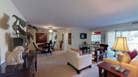 This thoughtfully designed single-floor residence offers a seamless blend of comfort and functionality across its well-proportioned living spaces. The open-concept living and dining area forms the heart of the home, where plush carpeting, ceiling fans, and neutral wall tones create a warm, inviting atmosphere. The living room benefits from a large sliding glass door that welcomes abundant natural light and provides access to an outdoor balcony, while framed wall art and tasteful décor enhance the overall aesthetic.

The dining area features a mirrored wall accent that amplifies the sense of space, and connects directly to a galley-style kitchen outfitted with rich dark wood cabinetry, granite-style countertops, tile flooring, and a full suite of stainless steel and black appliances including a gas range, refrigerator, and dishwasher.

The bedroom is generously sized with soft carpeting, a walk-in closet, and a window that fills the room with natural light. Two bathrooms serve the residence, each featuring white countertop vanities with undermount sinks, dark wood cabinetry, brushed nickel fixtures, and large mirrors with built-in medicine cabinets. The overall layout, as seen in the dollhouse view, is efficient and well-organized, making excellent use of every area throughout the home.