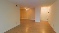 This single-floor residence offers a straightforward and practical layout designed for comfortable everyday living. The open living area is a defining feature of the space, showcasing warm hardwood flooring that runs throughout and lends a cohesive, inviting feel to the interior. Neutral-toned walls in a soft beige palette complement the natural warmth of the floors, creating a bright and versatile backdrop suited to a variety of interior styles. A ceiling fan with a built-in light fixture is centrally positioned in the main living area, providing both air circulation and ambient lighting. The space benefits from a partially open floor plan, with a structural column subtly defining the transition between living zones without fully enclosing them, maintaining a sense of openness. Recessed ceiling vents indicate a central HVAC system for year-round climate control. White baseboards provide a clean contrast against the flooring and walls, adding a polished finishing touch throughout. The overall ambiance is calm and move-in ready, offering a blank canvas well-suited for personalization. The layout is functional and efficient, making it an accessible and practical living space for a range of occupants.