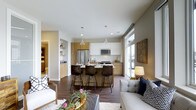 This single-floor space showcases a thoughtfully designed open-concept layout that seamlessly integrates the kitchen, dining, and living areas into one cohesive environment. The kitchen is a standout feature, outfitted with contemporary two-tone cabinetry — combining warm wood-toned lower cabinets with crisp white upper cabinets — and stainless steel appliances including a refrigerator, microwave, and range. A central island with a light stone countertop serves as both a prep space and a casual dining area, accommodated by sleek dark barstools. Pendant lighting above the island adds a refined touch. Rich, dark hardwood flooring runs throughout the space, grounding the design with warmth and continuity. The living area is bathed in natural light, courtesy of large windows that allow ample daylight to fill the room. Built-in shelving in an adjacent nook provides functional display and storage space. The overall ambiance is clean, modern, and inviting — striking a balance between contemporary design and comfortable livability, making it well-suited for both everyday living and entertaining.