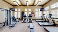 Located at 12302 S Yukon Ave in Glenpool, Oklahoma, this fitness center offers a well-equipped exercise space designed to support a full range of workout routines. The single-floor facility features soaring ceilings with distinctive dark wood beam accents, lending the room a refined architectural character. Warm, neutral-toned walls are complemented by durable striped carpet flooring throughout.

Natural light pours in through a generous bank of large windows fitted with white horizontal blinds, creating a bright and energizing atmosphere. Elevated transom windows further enhance the sense of openness and height. Overhead, flush-mounted rectangular lighting fixtures provide consistent illumination throughout the space.

The room is outfitted with a comprehensive selection of fitness equipment, including multiple TRUE-brand treadmills with integrated displays, elliptical cross-trainers, and a recumbent stationary bike. Strength training is supported by a multi-function cable machine and a flat workout bench. Free weight options include a rack of medicine balls in varying weights, stability/exercise balls, and a set of dumbbells. Wall-mounted flat-screen televisions provide entertainment during workouts. A community bulletin board and sanitizing station are also present. French-style glass doors and a solid wood entry door provide access to and from the adjoining corridor.