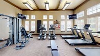 Located at 12302 S Yukon Ave in Glenpool, Oklahoma, this fitness center offers a well-equipped exercise space designed to support a full range of workout routines. The single-floor facility features soaring ceilings with distinctive dark wood beam accents, lending the room a refined architectural character. Warm, neutral-toned walls are complemented by durable striped carpet flooring throughout.

Natural light pours in through a generous bank of large windows fitted with white horizontal blinds, creating a bright and energizing atmosphere. Elevated transom windows further enhance the sense of openness and height. Overhead, flush-mounted rectangular lighting fixtures provide consistent illumination throughout the space.

The room is outfitted with a comprehensive selection of fitness equipment, including multiple TRUE-brand treadmills with integrated displays, elliptical cross-trainers, and a recumbent stationary bike. Strength training is supported by a multi-function cable machine and a flat workout bench. Free weight options include a rack of medicine balls in varying weights, stability/exercise balls, and a set of dumbbells. Wall-mounted flat-screen televisions provide entertainment during workouts. A community bulletin board and sanitizing station are also present. French-style glass doors and a solid wood entry door provide access to and from the adjoining corridor.