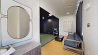 This single-floor indoor golf simulator space is thoughtfully designed for both practice and entertainment. The room is divided into two functional zones: a dedicated simulator bay and an adjacent lounge area. The simulator bay features a lush artificial turf floor, matte black walls that minimize glare and enhance the visual experience, and a large projection screen powered by a TruGolf simulation system. The system's computer and hardware stack are neatly housed on a multi-tier rack, allowing players to access a variety of virtual courses and practice modes. A basket of golf balls is conveniently available at the tee position. The lounge area transitions seamlessly from the simulator bay, finished with warm light wood flooring and crisp white walls. It is furnished with a modern geometric-patterned sofa and a low coffee table, providing comfortable seating for spectators. Heavy charcoal blackout curtains divide the two areas and aid in controlling light levels during play. An entry door allows natural light into the lounge when open. Wall-mounted artwork adds a decorative touch, and a decorative mirror with a gold-toned frame is positioned near the entrance, contributing to the space's polished, contemporary aesthetic.