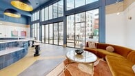 Located at 4101 Raleigh St in Charlotte, NC, this generously sized single-floor community lounge is a standout amenity space defined by its bold, contemporary design and thoughtfully curated interiors. Soaring double-height ceilings create an expansive, airy atmosphere, while an extensive wall of floor-to-ceiling windows and sliding glass doors floods the space with natural light and offers direct views and access to an outdoor courtyard area. The signature color palette of deep blue, warm yellow, and natural wood tones is carried throughout, lending the space a cohesive, energetic character. A striking curved island clad in deep blue vertical tile serves as a central focal point, topped with a white stone countertop and surrounded by sculptural matte black stools with white cushioned seats, beneath dramatic black-and-gold pendant lights. Built-in open shelving and cabinetry with stone countertops line one wall, styled with books, plants, and decorative objects. Cozy banquette-style seating with light wood frames and plush upholstery provides intimate gathering areas, complemented by lounge seating with warm-toned sofas and area rugs. A billiards table adds a recreational element, while curated wall art enhances the inviting ambiance throughout this well-appointed communal space.