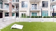Located at 6806 Morrison Blvd in Charlotte's SouthPark area, this single-level outdoor courtyard space offers a well-designed communal environment framed by a contemporary multi-story building. The architecture features a clean, modern aesthetic with neutral-toned paneling, large teal-framed windows, and curved balconies that define the courtyard's boundaries and provide a sense of enclosure without sacrificing openness.

The courtyard is thoughtfully divided into distinct functional zones. A stretch of vibrant artificial turf anchors the central area, serving as a dedicated lawn games space complete with a cornhole board. Strung bistro lights crisscross overhead, lending the space an inviting, social atmosphere during evening hours. Adjacent to the lawn, a paved deck area features Adirondack chairs and a small side table, creating an ideal spot for relaxed conversation or casual gatherings.

Raised concrete planters run along the perimeter, filled with a mix of shrubs, ornamental grasses, and small trees that introduce natural texture and greenery throughout. Paved walkways with a patterned tile design connect each zone, ensuring easy circulation. The overall ambiance is modern, polished, and well-suited for outdoor socializing in the heart of Charlotte.
