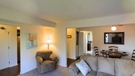 This single-floor residence offers a comfortable and well-organized layout designed for everyday living. The living area serves as the heart of the home, featuring plush wall-to-wall carpeting and neutral-toned walls that create a warm, welcoming atmosphere. The space is furnished with a generously sized sofa and a matching armchair, complemented by soft ambient lighting from a classic floor lamp that adds a cozy, relaxed feel to the room. A colorful floral artwork adorns one wall, adding a touch of personality to the understated decor. An open passage connects the living area to the rest of the home, offering a glimpse into adjoining spaces including what appears to be a hallway leading to additional rooms. The corridor beyond reveals doors that suggest a well-connected floor plan with private spaces tucked away from the main living zone. A decorative side table and chair are visible in the background, hinting at a transitional dining or entryway area. The overall ambiance is calm and residential, with consistent neutral tones throughout that lend the space a cohesive and uncluttered appearance. This single-story layout prioritizes ease of movement and accessibility, making it a practical and comfortable place to call home.