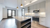 This contemporary single-floor residence showcases a thoughtfully designed open-concept layout with high-end finishes and a clean, modern aesthetic. The kitchen serves as the heart of the space, anchored by a substantial center island topped with striking granite countertops and accented by a duo of elegant gold pendant lights overhead. Soft gray shaker-style cabinetry extends floor to ceiling, complemented by a white subway tile backsplash and a full suite of stainless steel appliances, including a refrigerator, range, microwave, and dishwasher. Warm hardwood flooring flows seamlessly throughout the entire residence, unifying the living areas and bedrooms with a cohesive sense of warmth. The bedrooms are bright and well-proportioned, with large windows dressed with blinds that invite natural light while offering privacy, and bold dark-trimmed doors and baseboards providing sharp contrast against the crisp white walls. One bedroom features a set of black-framed French doors that open to an outdoor view, lending an airy, connected feel. The bathroom is finished with classic white tile, a tub-shower combination, and a clean vanity. Throughout the space, dark door frames and bold baseboards create a striking visual contrast, lending the residence a polished, contemporary character.