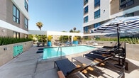 This well-appointed outdoor amenity space is designed with a resort-inspired aesthetic, offering a comprehensive range of leisure and recreation features set against the backdrop of a contemporary multi-story building. At the heart of the space is a sparkling swimming pool with clear turquoise water, surrounded by a generous sun deck outfitted with sleek, dark-framed chaise lounges and navy cushions. Striped blue-and-white umbrellas provide shaded seating areas furnished with lounge chairs and accent tables, creating comfortable spaces to relax poolside.

The outdoor kitchen area is a standout feature, equipped with a stainless steel built-in gas grill, granite countertops, a wet bar with seating, and decorative blue hexagonal tile accents — ideal for outdoor entertaining. Raised planter beds with ornamental grasses add greenery and natural texture throughout the deck, while the clean concrete paving unifies the entire outdoor floor plan.

An accessible indoor shower facility is also part of the amenity level, featuring bold teal patterned wall tiles, penny-tile flooring, and a grab bar, reflecting both thoughtful design and practical accessibility. Palm trees punctuate the perimeter, enhancing the warm, California-inspired atmosphere of this outdoor retreat.