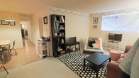 This single-floor space centers around a casual, functional living area with a warm, lived-in aesthetic. The room features light-colored walls that create a bright, open feel, complemented by natural light filtering in through a window along one wall. The flooring transitions between hardwood-style planks and a carpeted or neutral-toned area rug, which anchors the main seating zone and adds texture underfoot.

A tall, dark bookshelf serves as a focal point, filled with books, decorative items, and personal mementos, providing both storage and visual interest. Nearby, a compact media console holds a flat-screen television, making this a clear entertainment hub. Decorative string lights are draped along the upper perimeter of the walls, lending a warm, ambient glow to the space.

The seating area features a futon-style sofa dressed with patterned throw pillows in black, white, and orange, positioned beside the window. A bold black-and-white polka dot area rug defines the living zone, paired with a low black coffee table. Along the adjacent wall, a white shelving unit provides additional storage. Overall, the space feels cozy, personalized, and efficiently arranged.