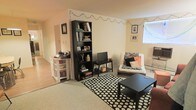 This single-floor space centers around a casual, functional living area with a warm, lived-in aesthetic. The room features light-colored walls that create a bright, open feel, complemented by natural light filtering in through a window along one wall. The flooring transitions between hardwood-style planks and a carpeted or neutral-toned area rug, which anchors the main seating zone and adds texture underfoot.

A tall, dark bookshelf serves as a focal point, filled with books, decorative items, and personal mementos, providing both storage and visual interest. Nearby, a compact media console holds a flat-screen television, making this a clear entertainment hub. Decorative string lights are draped along the upper perimeter of the walls, lending a warm, ambient glow to the space.

The seating area features a futon-style sofa dressed with patterned throw pillows in black, white, and orange, positioned beside the window. A bold black-and-white polka dot area rug defines the living zone, paired with a low black coffee table. Along the adjacent wall, a white shelving unit provides additional storage. Overall, the space feels cozy, personalized, and efficiently arranged.