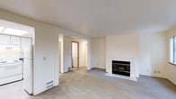 This single-floor residence offers a clean, straightforward layout designed for comfortable everyday living. The main living area is finished with wall-to-wall carpet in a neutral gray tone, creating a warm and understated foundation for the space. A standout feature of this room is the fireplace, framed by a white tile surround and hearth, which serves as a natural focal point along the wall. The room is bathed in a soft, even light, enhanced by the cream-colored walls and white ceiling that keep the space feeling bright and open. Floor vents are positioned throughout, indicating a forced-air heating and cooling system integrated seamlessly into the design. An open passageway leads from the living area into a hallway that connects to additional rooms, providing a sense of flow and accessibility throughout the home. The hallway reveals carpeted continuation and access to further interior spaces, including what appears to be a closet or utility door. The overall aesthetic is minimal and neutral, offering a versatile canvas well-suited to a variety of interior styles. The space is move-in ready and presents a practical, well-maintained environment ideal for residential living.