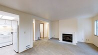 This single-floor residence offers a clean, straightforward layout designed for comfortable everyday living. The main living area is finished with wall-to-wall carpet in a neutral gray tone, creating a warm and understated foundation for the space. A standout feature of this room is the fireplace, framed by a white tile surround and hearth, which serves as a natural focal point along the wall. The room is bathed in a soft, even light, enhanced by the cream-colored walls and white ceiling that keep the space feeling bright and open. Floor vents are positioned throughout, indicating a forced-air heating and cooling system integrated seamlessly into the design. An open passageway leads from the living area into a hallway that connects to additional rooms, providing a sense of flow and accessibility throughout the home. The hallway reveals carpeted continuation and access to further interior spaces, including what appears to be a closet or utility door. The overall aesthetic is minimal and neutral, offering a versatile canvas well-suited to a variety of interior styles. The space is move-in ready and presents a practical, well-maintained environment ideal for residential living.