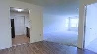 This single-floor residence offers a straightforward and functional layout designed for comfortable everyday living. The main living area is generously proportioned, featuring wall-to-wall carpeting that adds warmth underfoot. Large windows allow abundant natural light to pour into the space, creating a bright and airy atmosphere. The neutral, light-toned walls throughout provide a clean and versatile backdrop.

A distinct transition in flooring marks the entry into the kitchen area, where durable hardwood-style planks replace the carpeting. The kitchen is a separate but adjacent space, visible through an open doorway, and is equipped with standard appliances including a range. Cabinetry in a dark wood finish provides contrast against the lighter walls. A bifold-door closet is also visible nearby, offering convenient storage.

The open flow between the living and kitchen zones makes the space feel connected while maintaining a clear delineation between functional areas. The overall ambiance is clean, neutral, and move-in ready, with simple finishes that offer flexibility for personalization. The single-floor plan ensures easy accessibility and a cohesive living experience throughout.