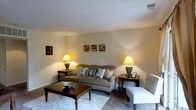 This single-floor space presents a comfortable and well-appointed living area designed with a warm, welcoming ambiance. The living room serves as the focal point, featuring neutral-toned walls that lend a soft, airy feel to the interior. A textured ceiling adds subtle architectural character to the room.

The seating arrangement centers around a classic upholstered sofa adorned with decorative teal and patterned throw pillows, complemented by a coordinating accent chair. A dark wood coffee table with turned legs anchors the seating area, topped with a decorative bowl as a stylish accent piece. A patterned area rug defines the space and adds visual interest to the carpeted floor.

Warm ambient lighting is provided by two matching table lamps positioned on dark wood end tables flanking the sofa, creating a balanced and symmetrical arrangement. A small collection of framed wall art, including abstract circular prints, adds a decorative touch above the sofa. The overall design aesthetic is traditionally inspired, offering a functional yet inviting environment suitable for relaxing or entertaining.