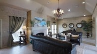 This single-floor space presents a thoughtfully designed interior that blends elegance with comfort. The main lounge area serves as the centerpiece, featuring a striking vaulted ceiling that adds a sense of grandeur and openness to the room. A decorative white fireplace with a detailed mantelpiece anchors one wall, topped with a vivid landscape painting depicting a sun-drenched Mediterranean coastal scene, lending an artistic focal point to the space.

The walls are finished in a warm, textured faux treatment in neutral earth tones, contributing to an inviting and refined ambiance. Rich dark leather sofas and upholstered seating are arranged to encourage conversation, complemented by ornate wooden accent tables. An elaborate chandelier with multi-arm candelabra styling hangs from the ceiling, providing warm, ambient lighting alongside recessed fixtures.

Natural light filters in through French doors along one wall, while decorative mirrors in round dark frames are mounted nearby, enhancing the sense of depth and light within the space. Tall decorative grasses and floral arrangements are thoughtfully placed throughout, adding organic texture. Tile flooring grounds the space with a clean, durable foundation. Overall, the space exudes a warm, upscale character with Mediterranean-inspired design sensibilities.