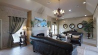 This single-floor space presents a thoughtfully designed interior that blends elegance with comfort. The main lounge area serves as the centerpiece, featuring a striking vaulted ceiling that adds a sense of grandeur and openness to the room. A decorative white fireplace with a detailed mantelpiece anchors one wall, topped with a vivid landscape painting depicting a sun-drenched Mediterranean coastal scene, lending an artistic focal point to the space.

The walls are finished in a warm, textured faux treatment in neutral earth tones, contributing to an inviting and refined ambiance. Rich dark leather sofas and upholstered seating are arranged to encourage conversation, complemented by ornate wooden accent tables. An elaborate chandelier with multi-arm candelabra styling hangs from the ceiling, providing warm, ambient lighting alongside recessed fixtures.

Natural light filters in through French doors along one wall, while decorative mirrors in round dark frames are mounted nearby, enhancing the sense of depth and light within the space. Tall decorative grasses and floral arrangements are thoughtfully placed throughout, adding organic texture. Tile flooring grounds the space with a clean, durable foundation. Overall, the space exudes a warm, upscale character with Mediterranean-inspired design sensibilities.