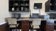 The business center at 4583251 is a single-floor facility thoughtfully designed to accommodate a range of professional needs. The space centers around a granite-topped built-in workstation that spans the width of the room, providing ample surface area for multiple users simultaneously. Three computer monitors are positioned along the counter, each paired with nailhead-trim upholstered bar-height chairs in a warm beige tone, combining functionality with a polished aesthetic. Dark espresso cabinetry with brushed nickel hardware is integrated beneath the countertop, offering convenient storage solutions. Decorative tall vases in black and teal add refined visual interest between the workstations. Above the desk area, open-cube wall shelving in a matching dark finish displays books and decorative accents, lending the space a cultivated, organized feel. A flat-screen television is mounted to the wall, adding a media resource to the environment. The overall palette of cream walls, dark wood tones, and granite surfaces creates a cohesive, professional ambiance suitable for focused work or business-related tasks.