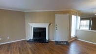 This single-floor space offers a welcoming and functional layout designed with classic interior details throughout. The main living area immediately draws attention with its rich, dark hardwood flooring that extends seamlessly across the room, adding warmth and depth to the overall aesthetic. A standout architectural feature is the traditional wood-burning fireplace, framed by an elegant white mantel with columned detailing and a dark tile hearth surround, serving as a natural focal point of the space. Crown molding runs along the ceiling perimeter, lending a refined, polished finish to the room. The walls are painted in a neutral warm beige tone, creating a versatile backdrop that complements both modern and traditional furnishing styles. A white interior entry door with standard hardware provides secure access, accompanied by a window that allows natural light to filter into the space. The clean, open layout of the living area offers ample flexibility for furniture arrangement. The combination of classic architectural elements, warm tones, and hardwood flooring gives the space a timeless, comfortable ambiance suitable for a variety of living arrangements.